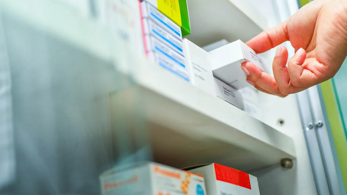 Hand reaching for a box of medication from a shelf in a pharmacy or medical storage unit.