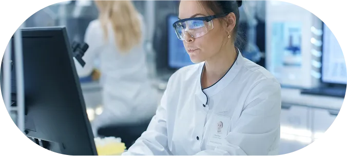 Female scientist wearing safety goggles and gloves working on a computer in a laboratory, with test tubes and lab equipment visible on the desk.