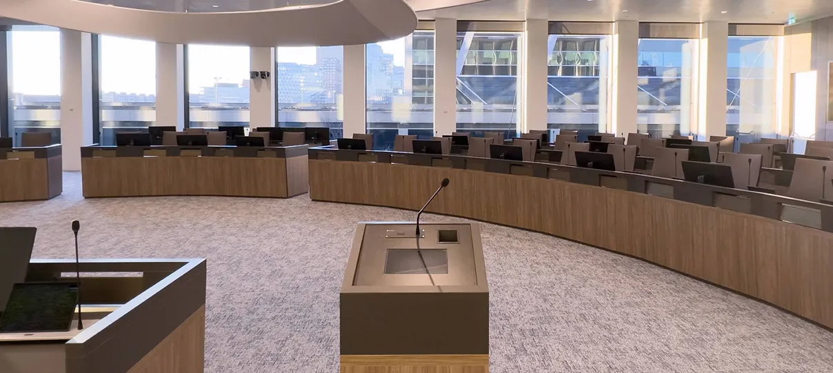 Modern conference or council chamber with a large U-shaped arrangement of desks, microphones, and screens, set in a room with tall windows and natural lighting.