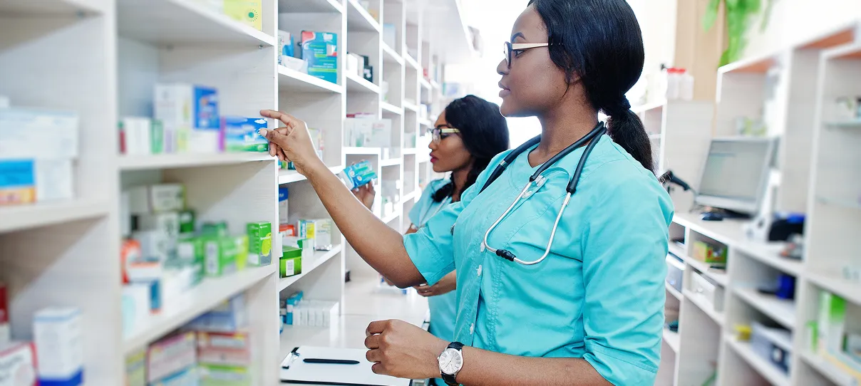 Two female pharmacists or healthcare workers in teal uniforms selecting medicine from shelves in a well-stocked pharmacy.