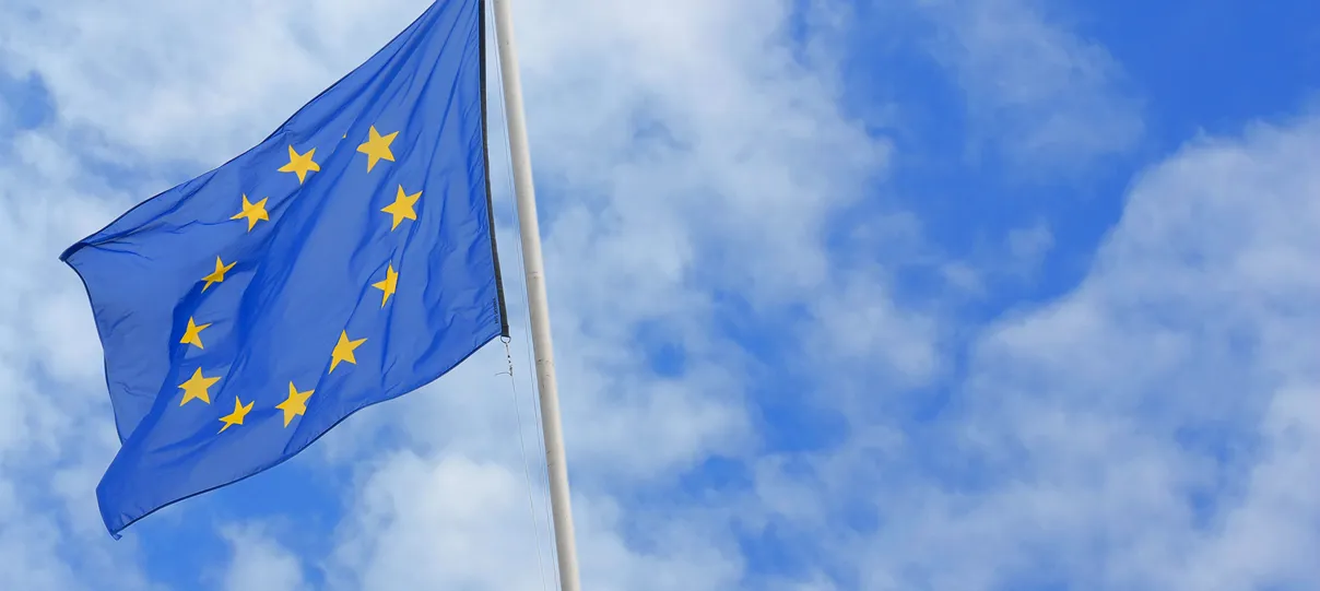European Union flag with yellow stars on a blue background waving on a flagpole against a partly cloudy sky.