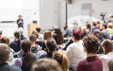 Audience seated in a lecture hall or conference room, listening to a speaker giving a presentation at the front of the room.