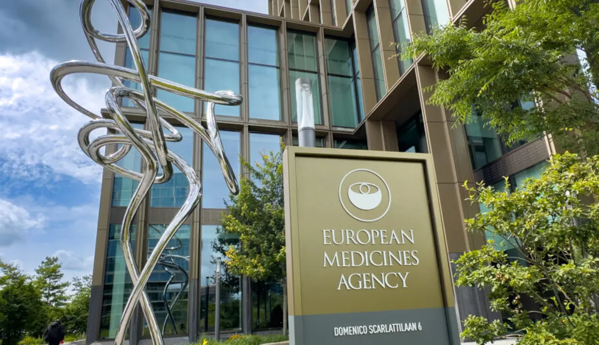 Modern conference or council chamber with a large U-shaped arrangement of desks, microphones, and screens, set in a room with tall windows and natural lighting.