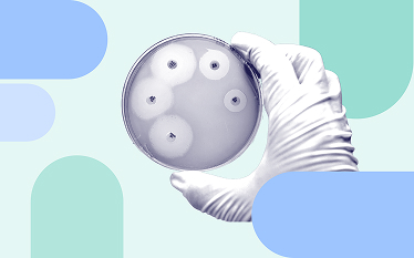 Gloved hand holding a petri dish with bacterial cultures or antibiotic testing spots, set against a stylised pastel background.