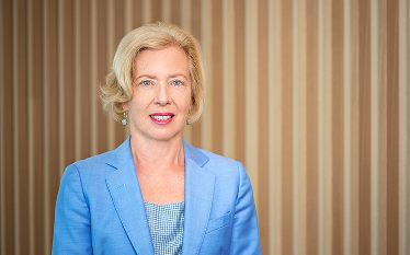 Portrait of a woman with blonde hair wearing a light blue blazer, standing in front of a background with vertical beige stripes.