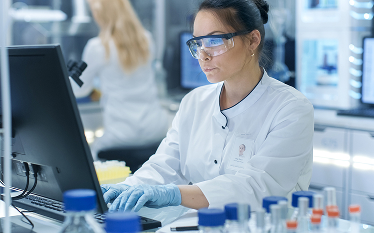 Female scientist wearing safety goggles and gloves working on a computer in a laboratory, with test tubes and lab equipment visible on the desk.