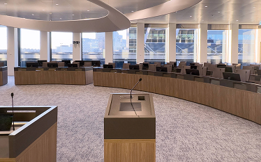 Modern conference or council chamber with a large U-shaped arrangement of desks, microphones, and screens, set in a room with tall windows and natural lighting.