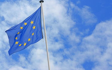 European Union flag with yellow stars on a blue background waving on a flagpole against a partly cloudy sky.