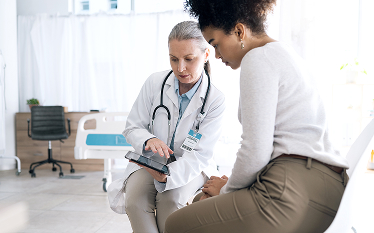 Female doctor showing medical information on a digital tablet to a patient in a hospital room, with a hospital bed and chair in the background.