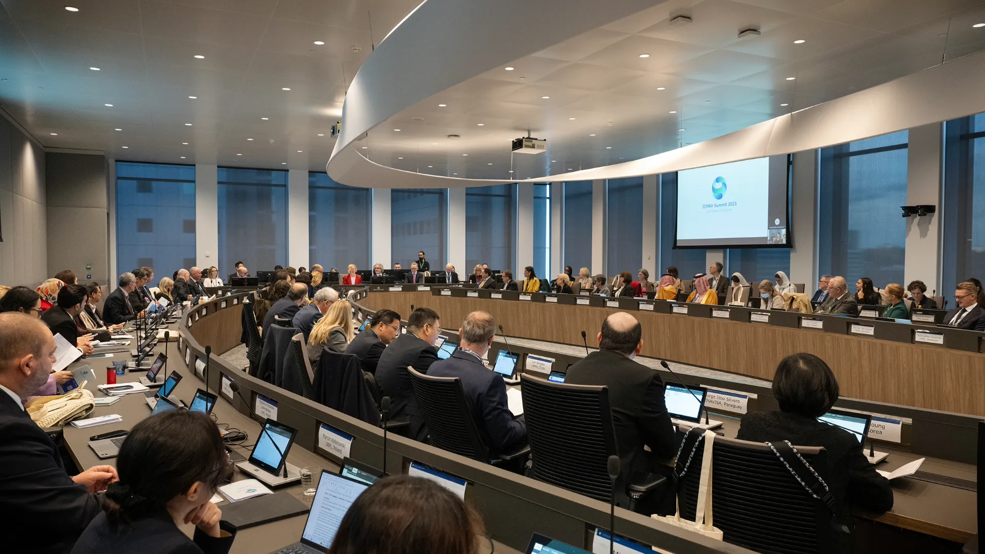 Modern conference or council chamber with a large U-shaped arrangement of desks, microphones, and screens, set in a room with tall windows and natural lighting.