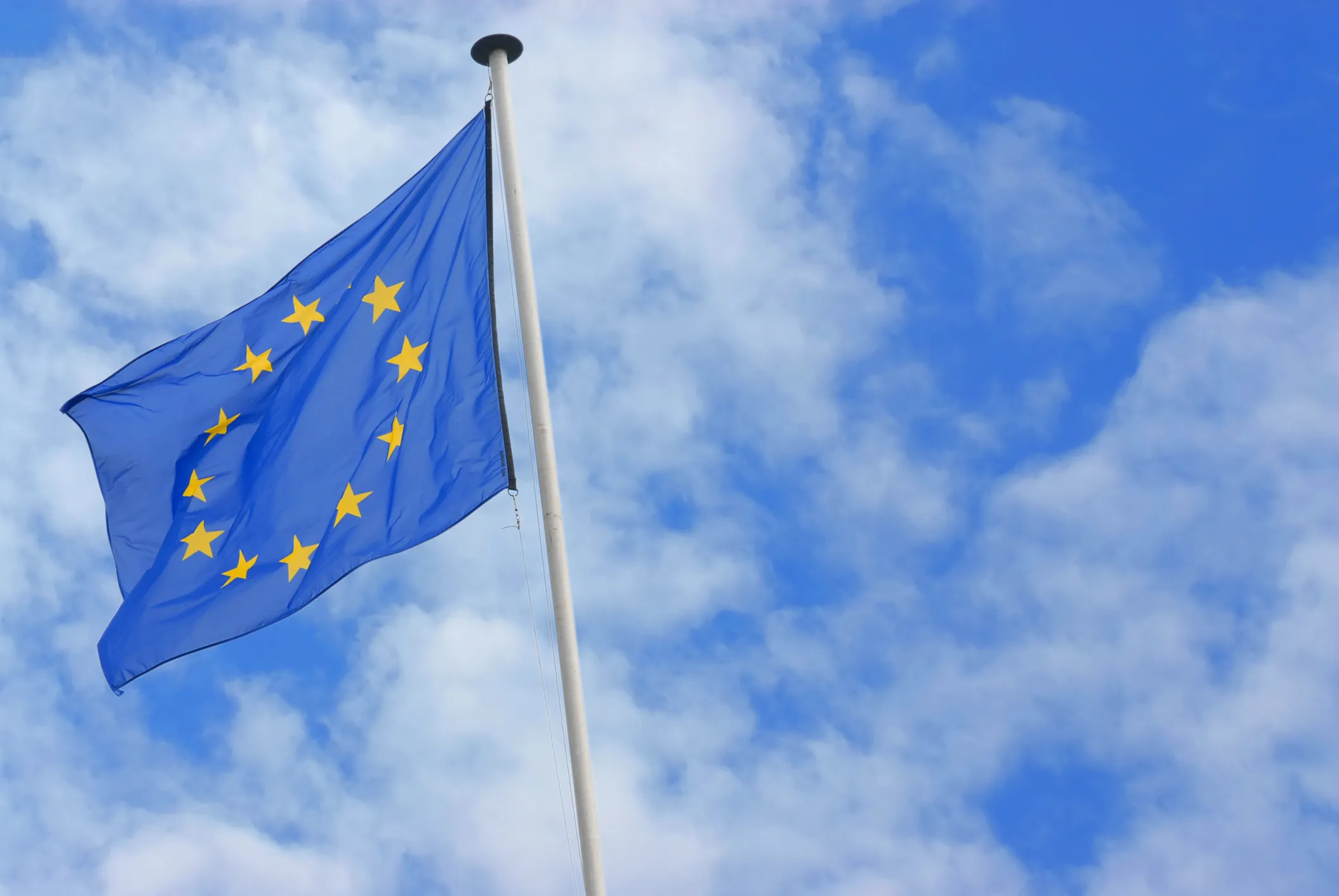 European Union flag with yellow stars on a blue background waving on a flagpole against a partly cloudy sky.