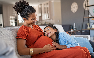 Pregnant person seated on a sofa with a child leaning against the abdomen.