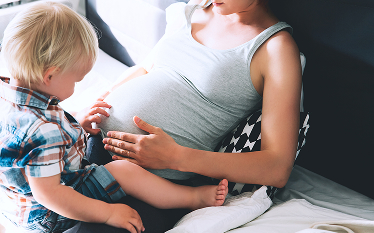 Pregnant person seated on a bed with a young child touching the abdomen