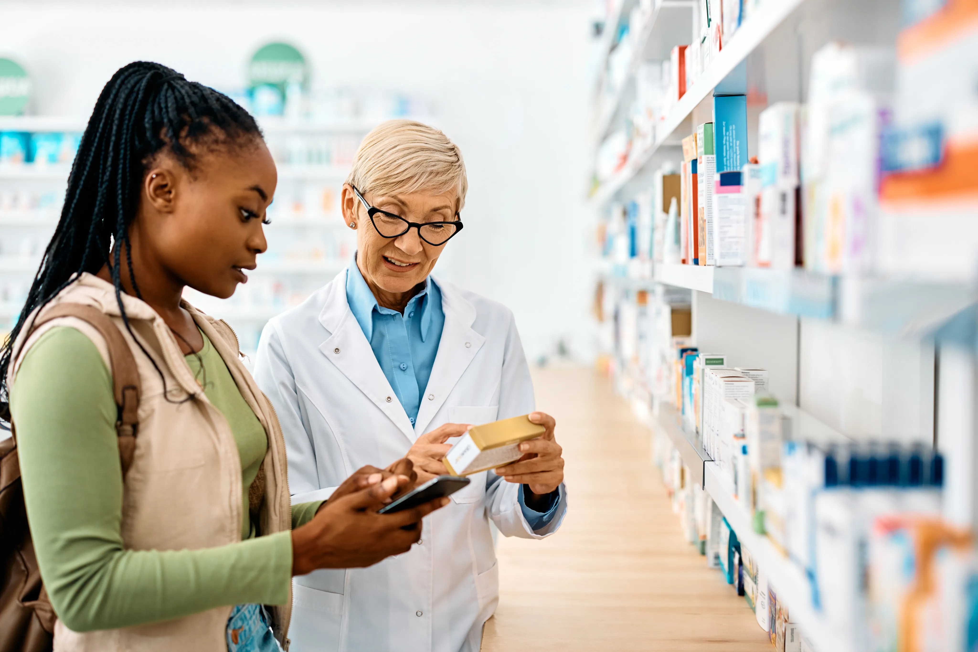 Two female pharmacists or healthcare workers in teal uniforms selecting medicine from shelves in a well-stocked pharmacy.