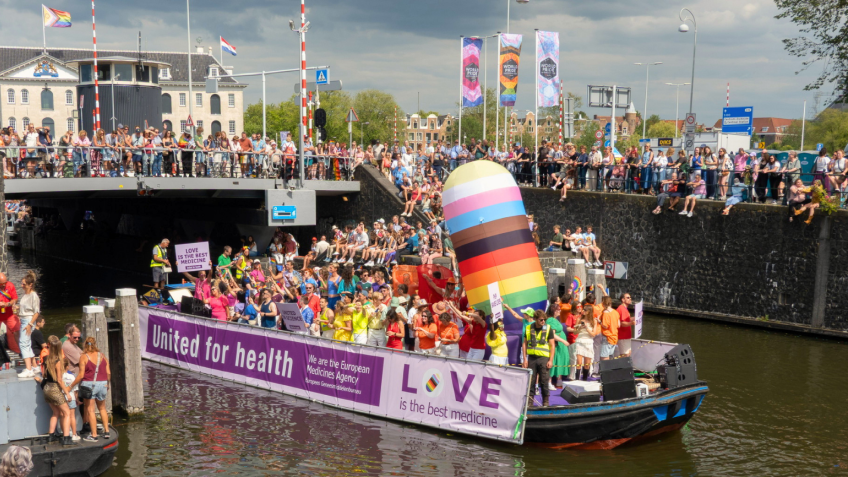 EMA's boat in the Worldpride 2026 Amsterdam canal parade displaying banners reading “United for health” and “LOVE is the best medicine,” with spectators lining the canal and bridge.