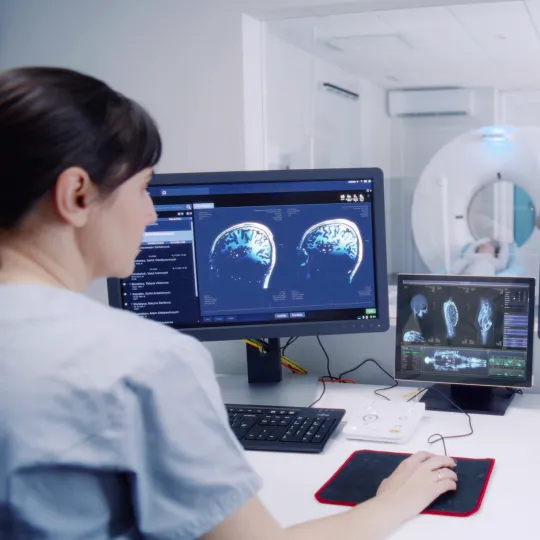 Medical professional at her desk looking at computer screens showing scans of a human body, in a room overlooking a scanning machine