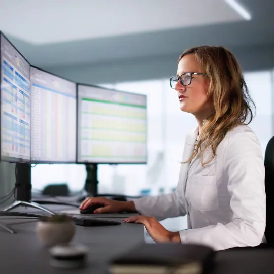 Person using two computer monitors displaying data tables and charts while typing at a desk in an office setting