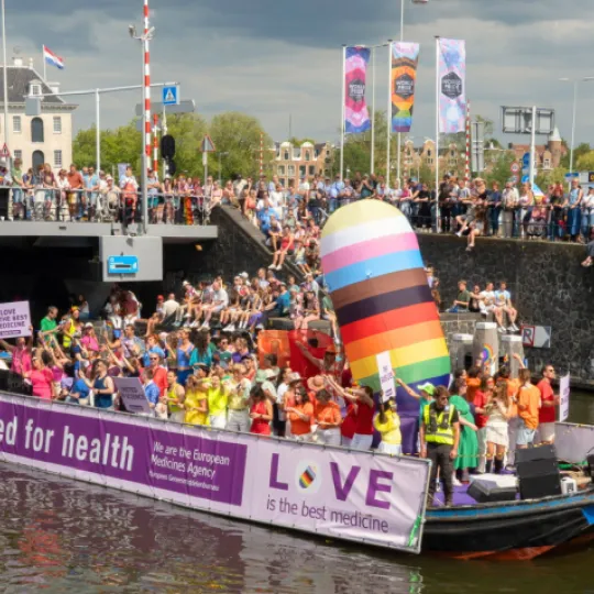 EMA's boat in the Worldpride 2026 Amsterdam canal parade displaying banners reading “United for health” and “LOVE is the best medicine,” with spectators lining the canal and bridge.